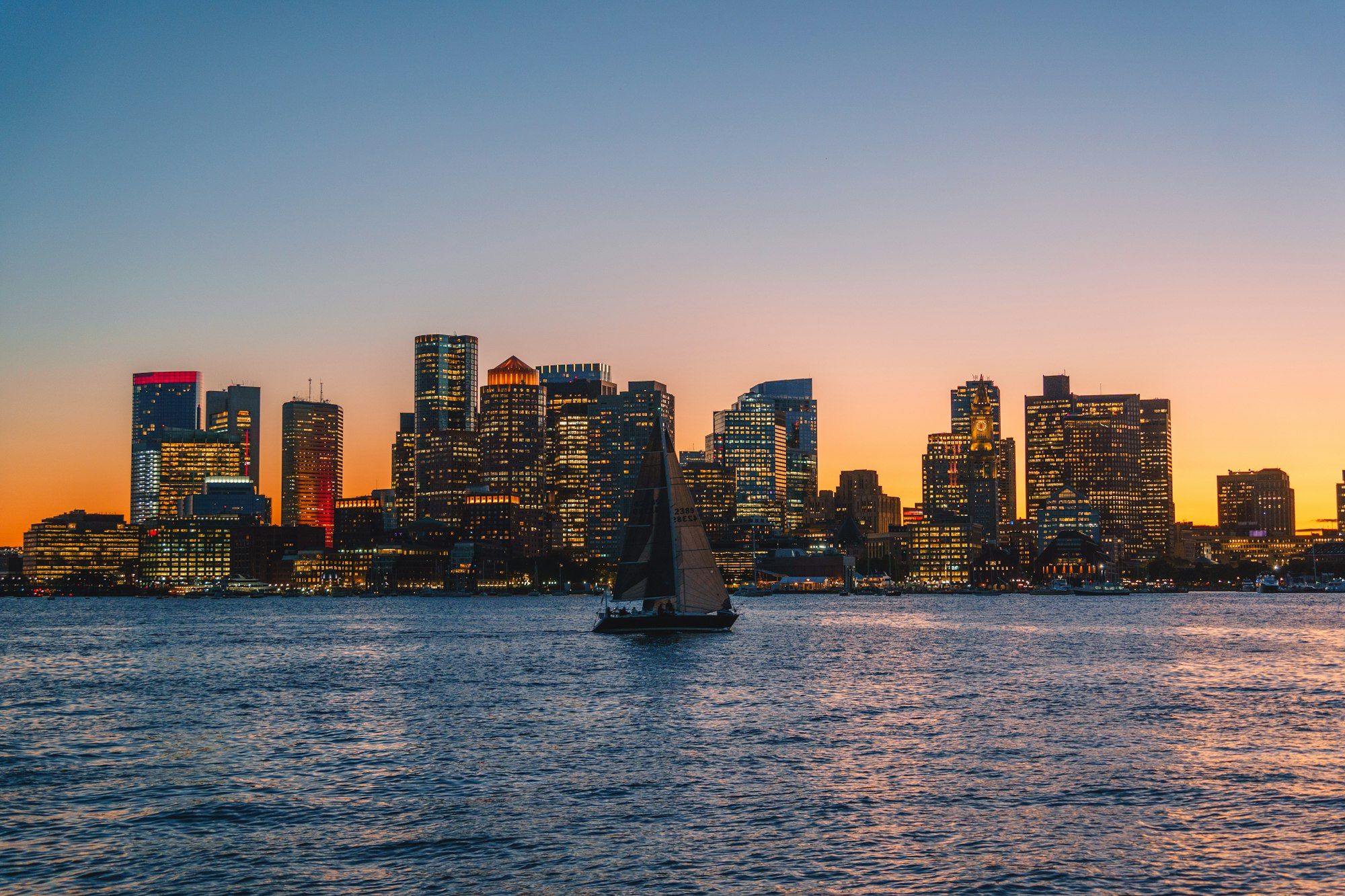 The Boston skyline across the harbor at sunset with a sailboat in the foreground