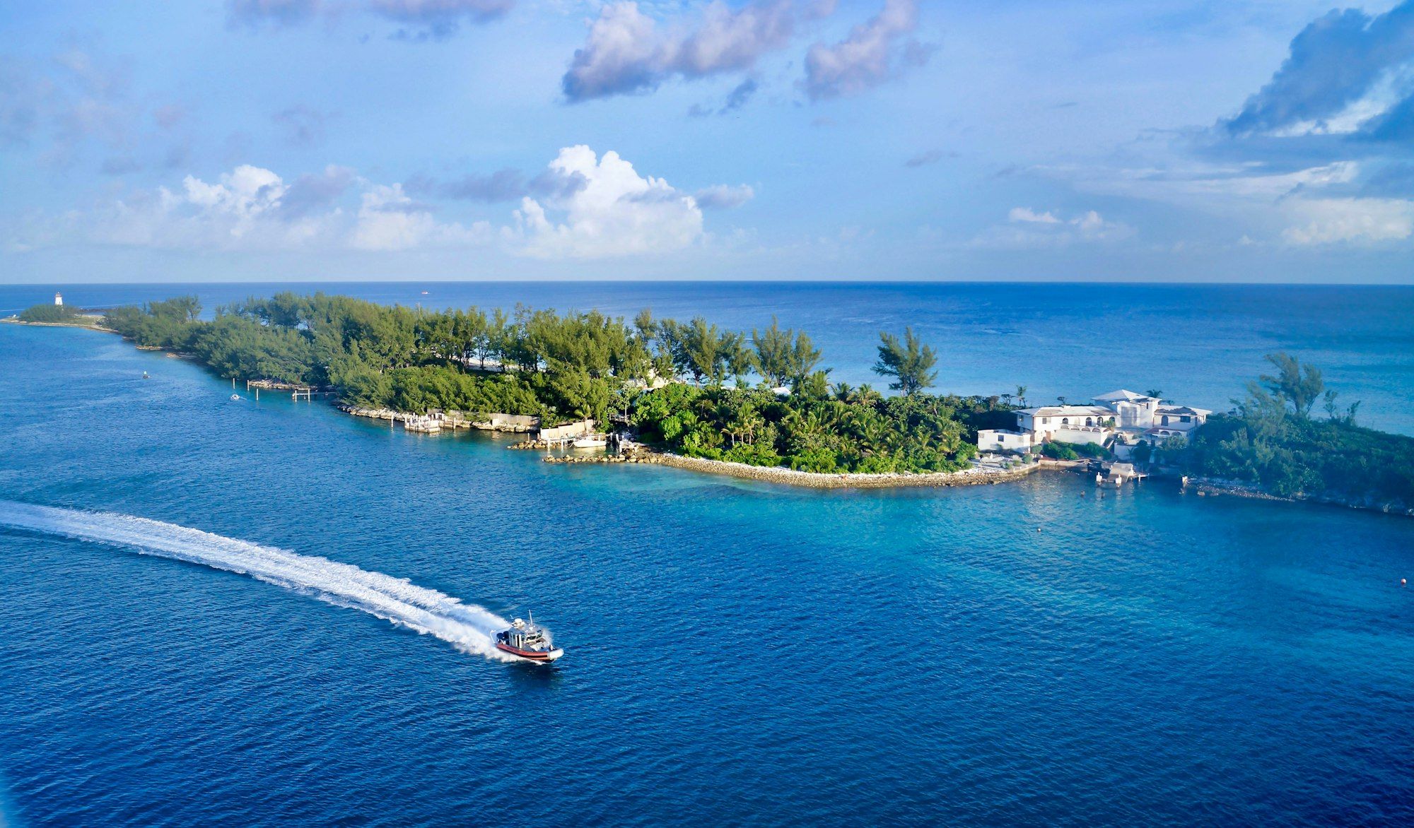 A patrol boat leaves a white wake off a wooded point at the entrance to Nassau harbour, with a lighthouse in the distance