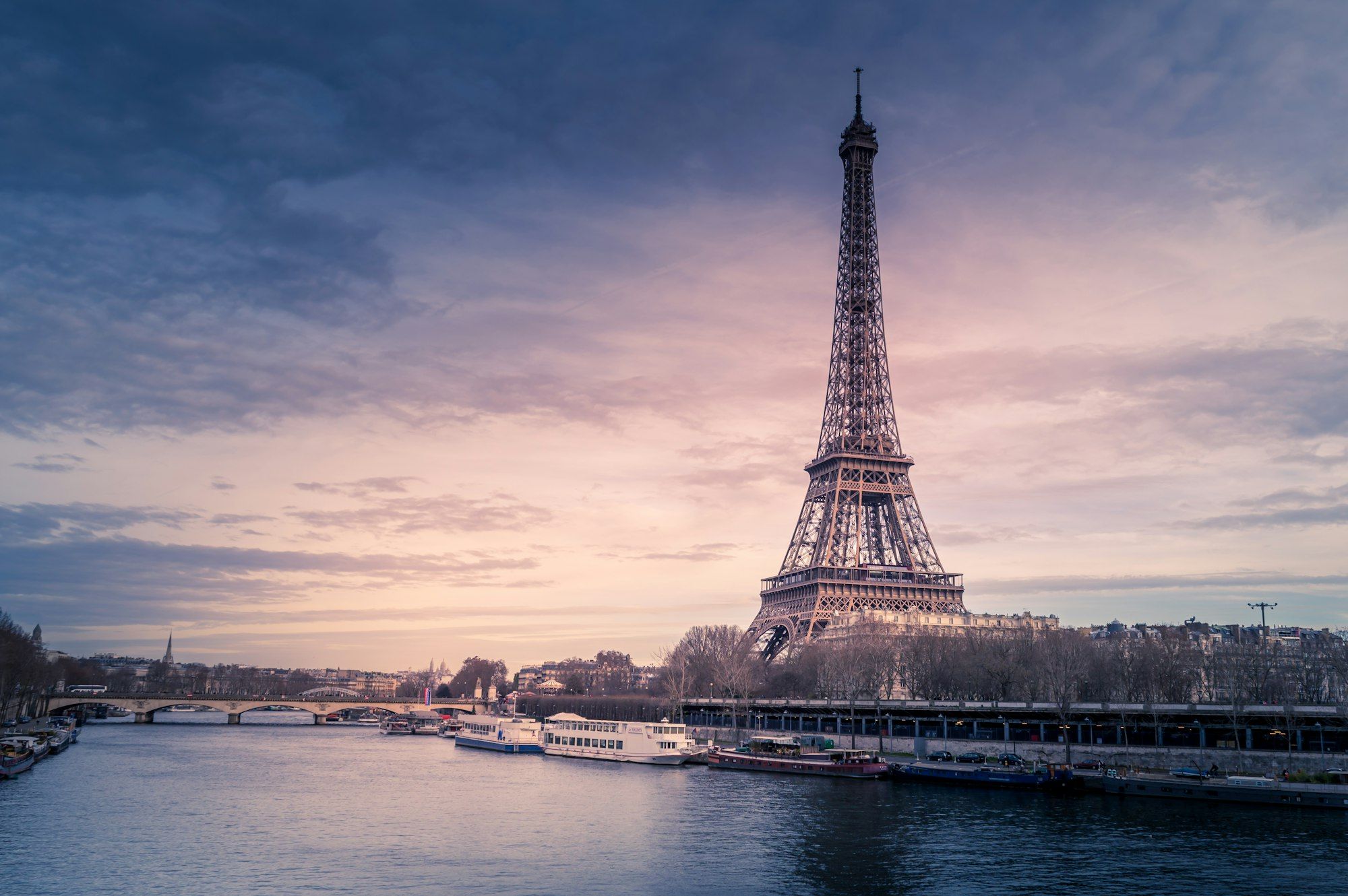 The Eiffel Tower rising above the Seine at dusk, with river boats moored along the quay